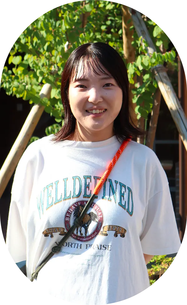 白いTシャツを着た女性が優しい笑顔で立っている。後ろには緑の木々が見える。