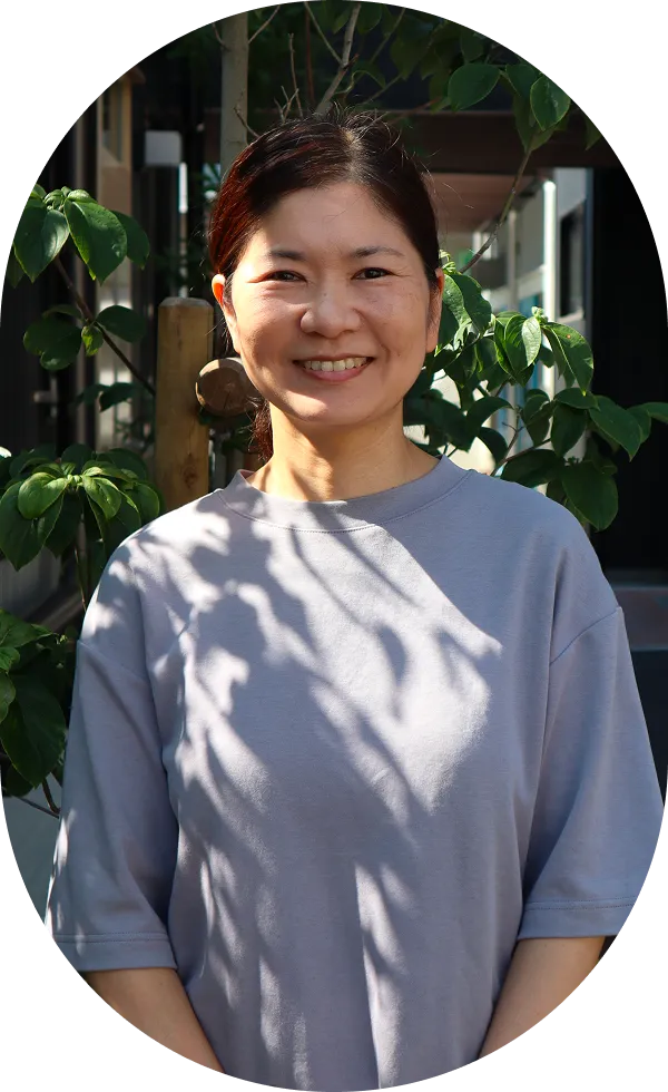 グレーのTシャツを着た女性が温かみのある笑顔で立っている。後ろには緑の木々が見える。