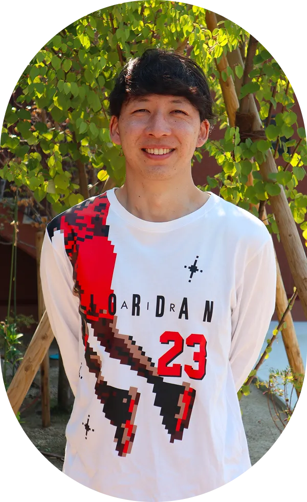 スポーティな柄の白いシャツを着た男性が爽やかな笑顔で立っている。後ろには緑の木々が見える。